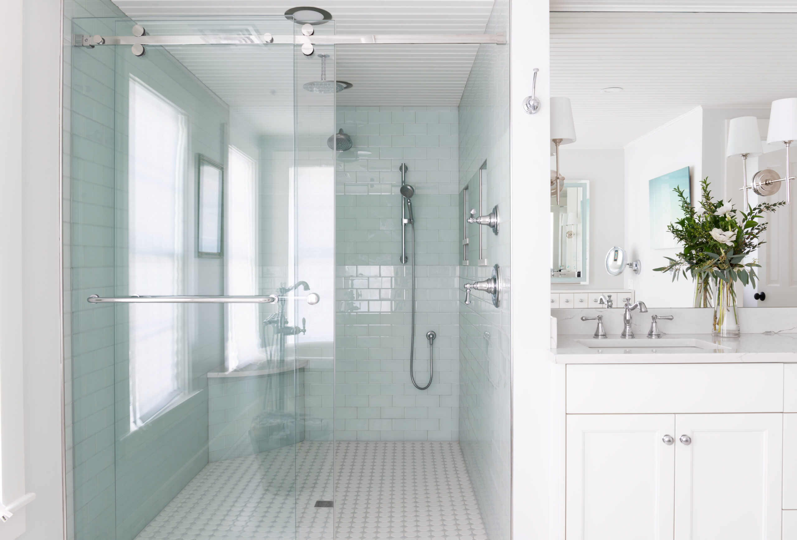 The image showcases a modern bathroom interior highlighting a spacious walk-in shower. The shower features pale blue subway tiles on the walls and small, hexagonal tiles on the floor, enhancing its clean and contemporary aesthetic. The glass enclosure provides a seamless look, complemented by chrome fixtures, including a large overhead shower head and a secondary hand-held unit. The bathroom is well-lit by natural light streaming through a frosted window, and the white vanity next to the shower is topped with a marble counter and equipped with stylish fixtures, providing a luxurious feel to the renovation process. This image is often used to illustrate the meticulous details and quality finishes involved in interior renovation projects.